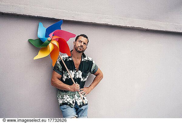 Thoughtful man with hand on hip holding pinwheel toy leaning on concrete wall