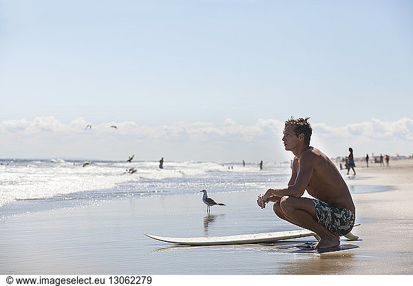 Thoughtful man with surfboard crouching at beach