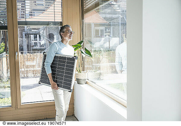 Thoughtful mature businessman standing with solar panel and looking through window