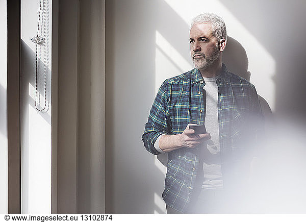 Thoughtful mature man holding smart phone looking through window at home