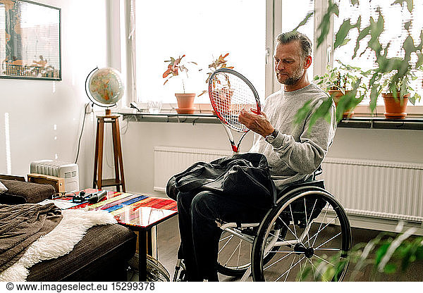 Thoughtful mature man holding tennis racket while sitting on wheelchair at home