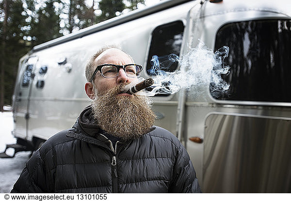 Thoughtful mature man smoking cigar outside caravan in forest