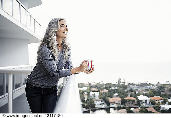 Thoughtful mature woman holding coffee mug while standing on balcony