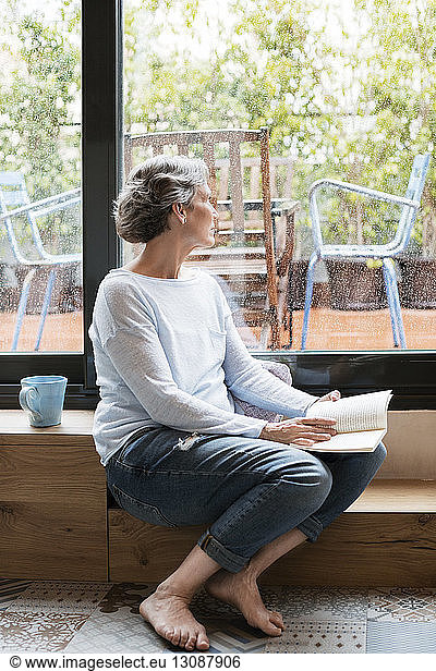 Thoughtful mature woman looking through window while holding book