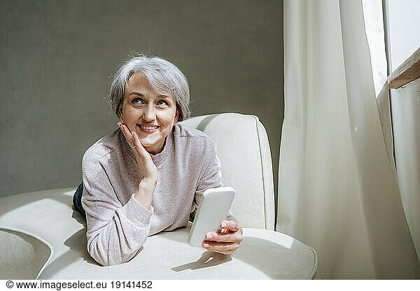 Thoughtful mature woman with smart phone on couch at home