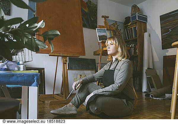 Thoughtful painter holding paintbrush sitting on floor in art studio