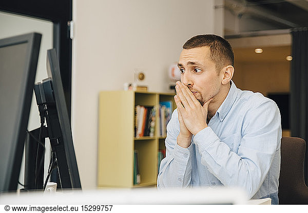 Thoughtful programmer with hands clasped looking at computer monitor while working at desk in office