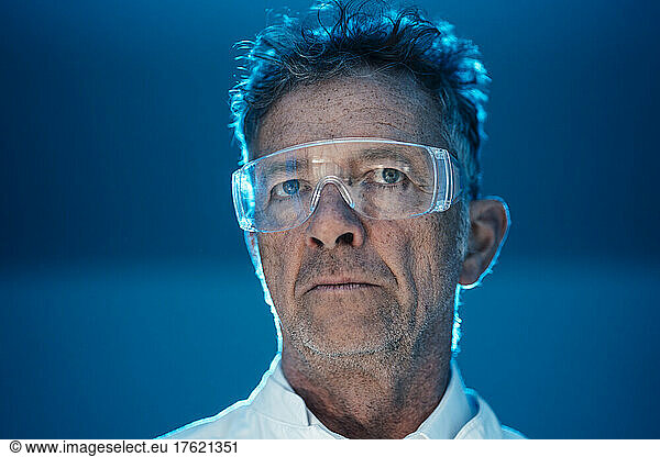 Thoughtful scientist with protective eyewear in front of blue wall