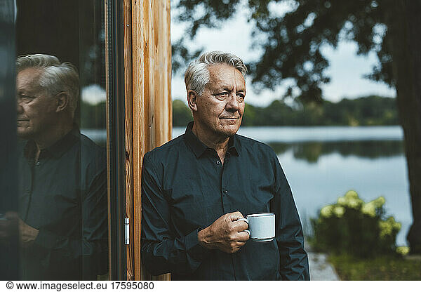 Thoughtful senior man holding coffee cup by glass wall at backyard