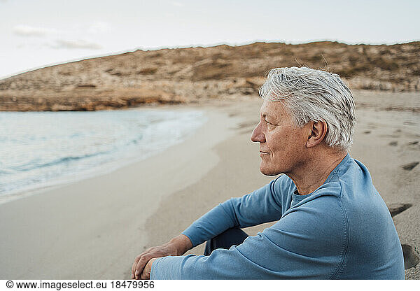 Thoughtful senior man sitting on beach at sunset