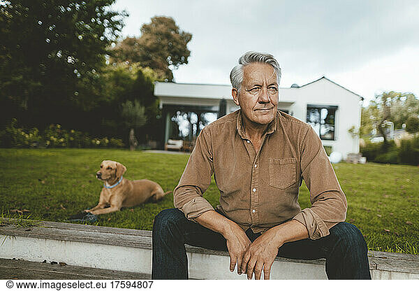 Thoughtful senior man sitting on steps by pet dog at backyard