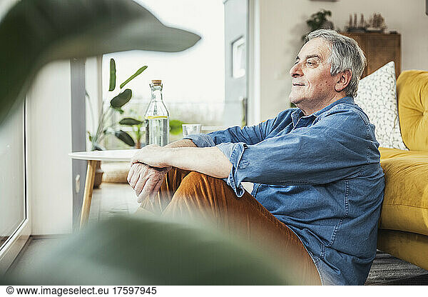 Thoughtful senior man with gray hair sitting by sofa in living room