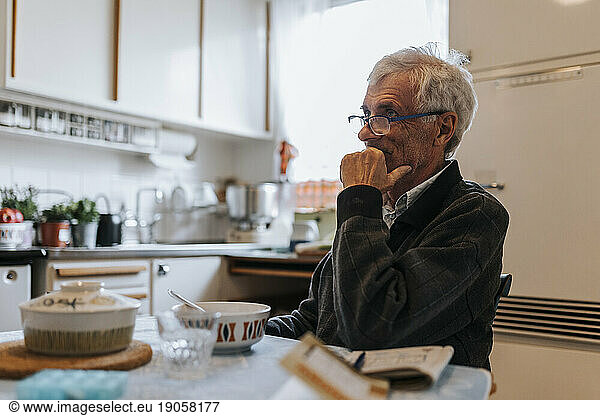 Thoughtful senior man with hand on chin sitting in kitchen at home