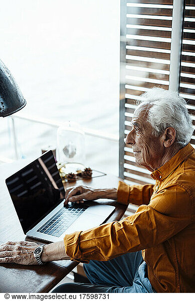 Thoughtful senior man with laptop sitting at table in houseboat