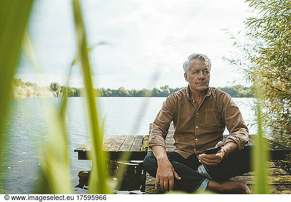 Thoughtful senior man with mobile phone sitting on jetty by lake