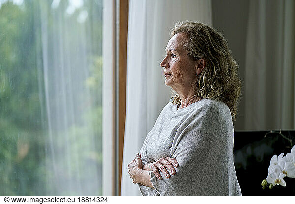 Thoughtful senior woman standing with arms crossed and looking through window at home