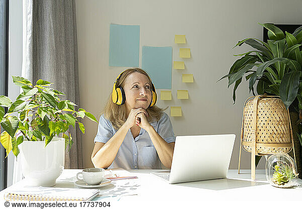 Thoughtful smiling freelancer listening music through wireless headphones sitting with laptop at desk in home office