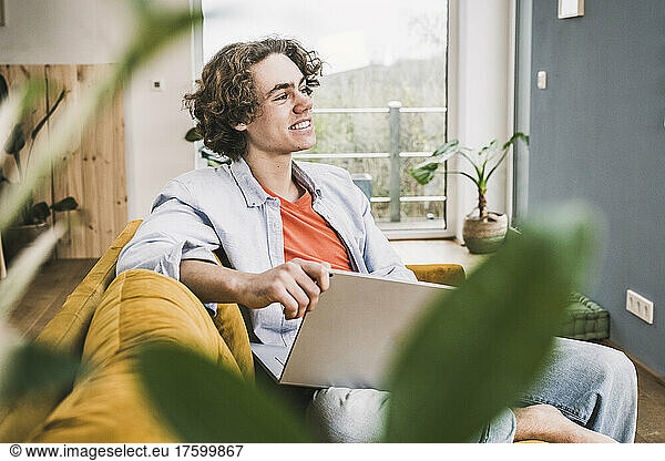 Thoughtful smiling man with laptop sitting on sofa in living room at home