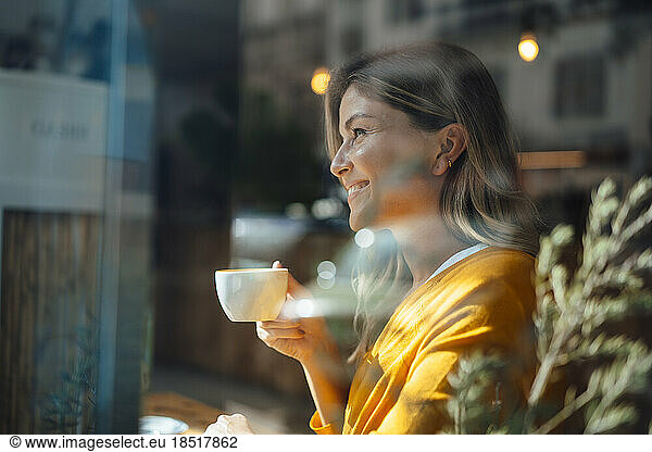 Thoughtful smiling woman having coffee in cafe seen through glass