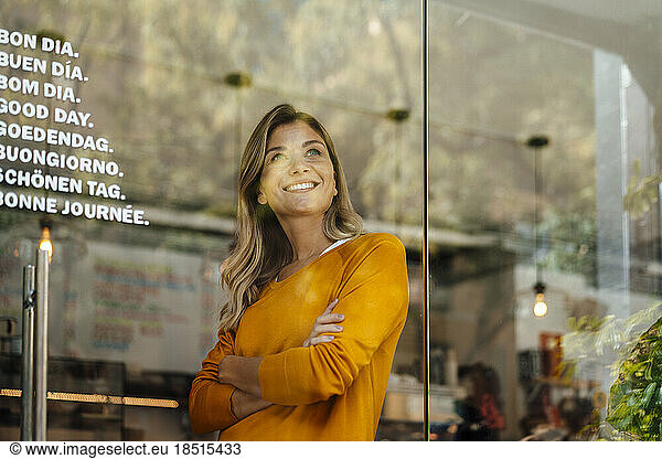 Thoughtful smiling woman with arms crossed in cafe seen through glass