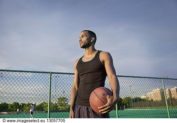 Thoughtful sportsman with basketball standing in court against sky