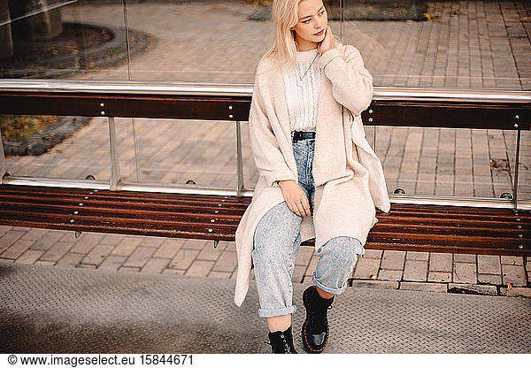 Thoughtful stylish young woman sitting on bus stop in city