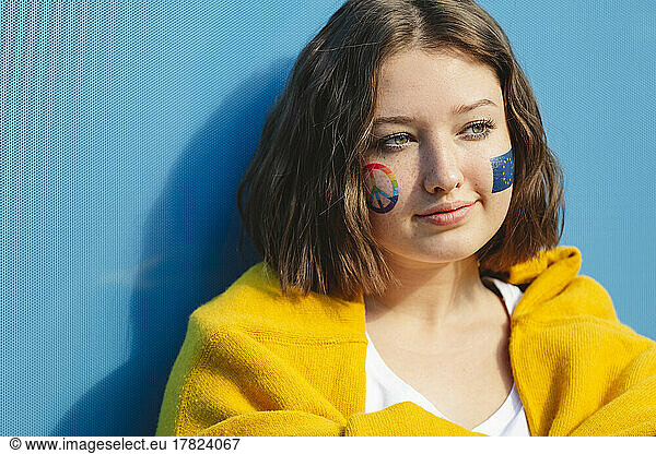 Thoughtful teenage girl with peace symbol and European Union paint on cheeks in front of blue wall