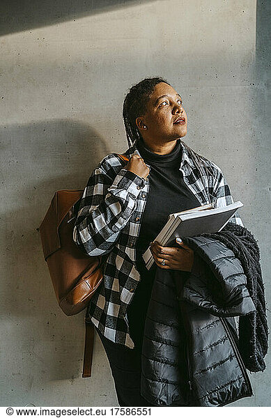 Thoughtful woman holding book and jacket standing with backpack against gray wall
