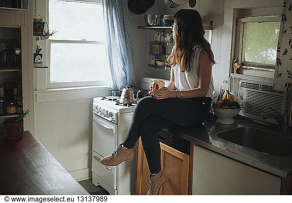 Thoughtful woman holding coffee cup while sitting on kitchen counter