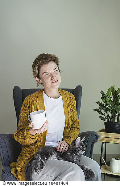 Thoughtful woman holding mug sitting with cat in armchair at home