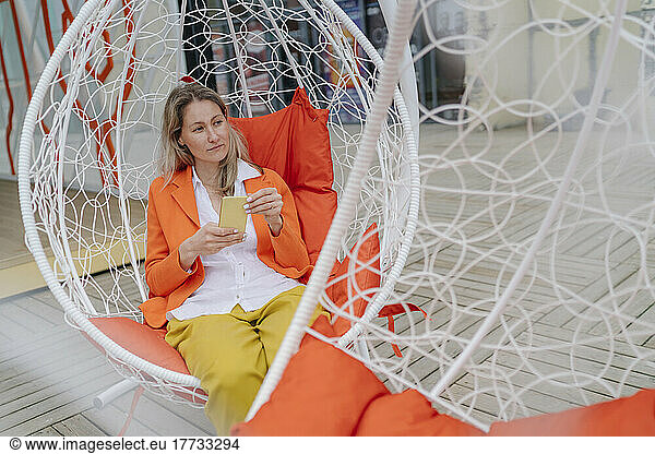 Thoughtful woman holding smart phone sitting on hanging chair