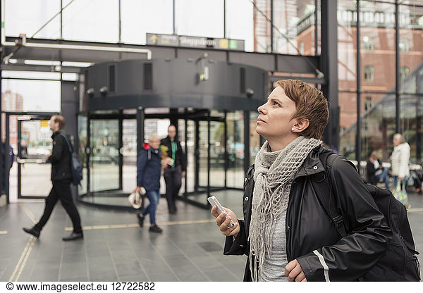 Thoughtful woman holding smart phone while looking up by building in city