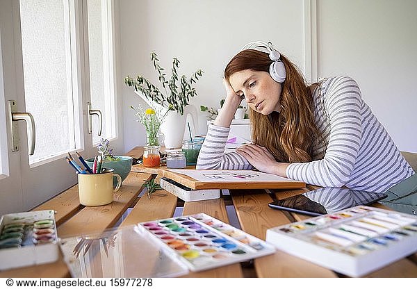 Thoughtful woman listening music though headphones while painting at home