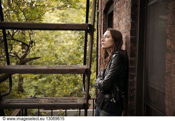 Thoughtful woman standing against wall by fire escape against trees