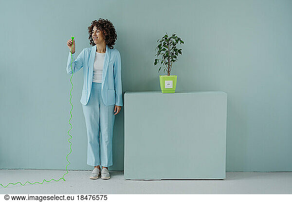 Thoughtful woman standing with electric plug by potted plant in front of wall