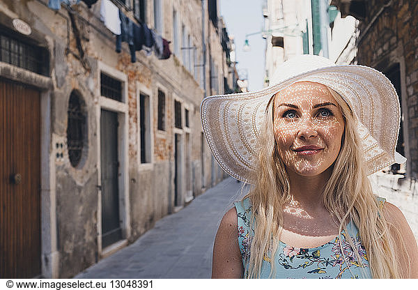 Thoughtful woman wearing hat while looking away at narrow footpath amidst buildings