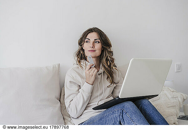Thoughtful woman with credit card and laptop sitting at home