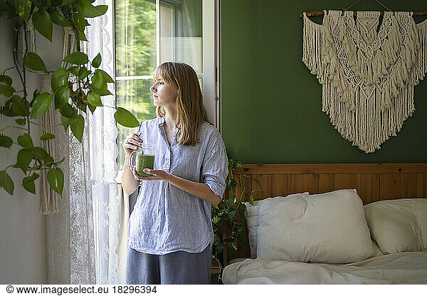 Thoughtful woman with green smoothie standing by window