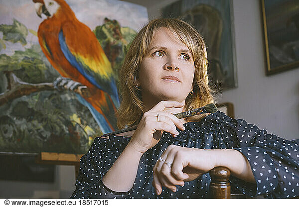 Thoughtful woman with hand on chin holding paintbrush in front of bird painting at workshop