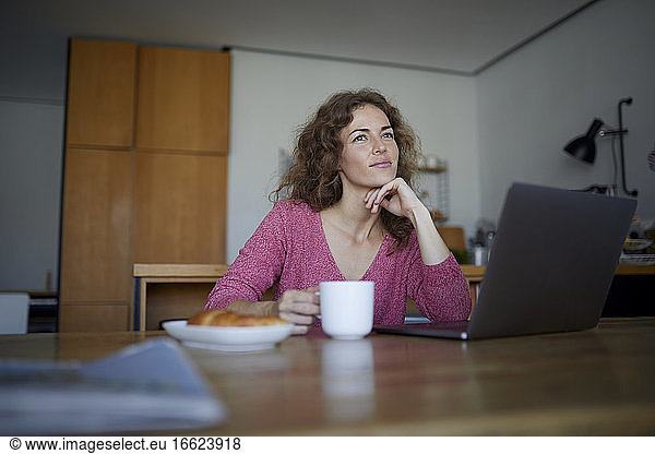 Thoughtful woman with laptop holding coffee cup while sitting at home