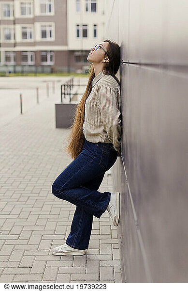 Thoughtful woman with long hair wearing eyeglasses leaning on wall