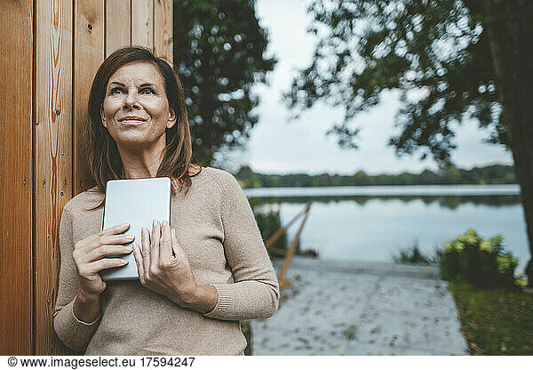 Thoughtful woman with tablet PC standing by wooden wall at backyard