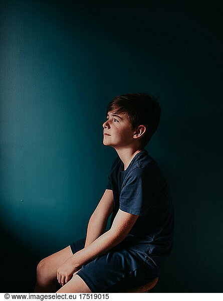 Thoughtful young boy sitting on a stool against a dark blue wall.
