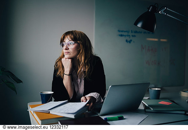 Thoughtful young businesswoman sitting with document and laptop at creative office