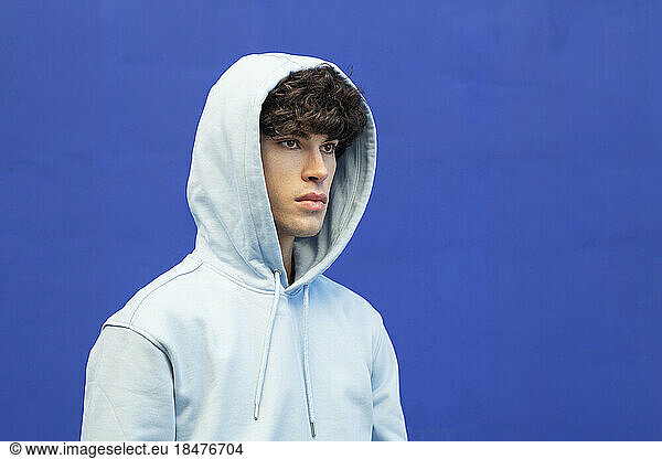 Thoughtful young man wearing hooded shirt in front of blue wall