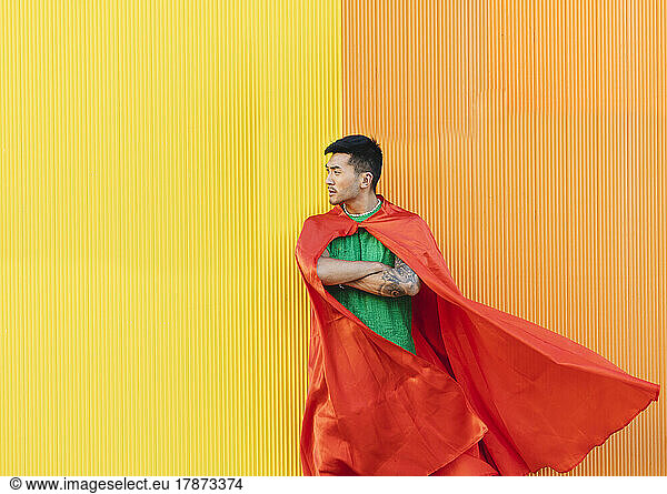 Thoughtful young man wearing red superhero cape standing in front of wall