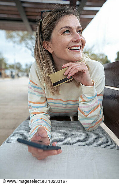 Thoughtful young woman lying on bench holding credit card and smart phone