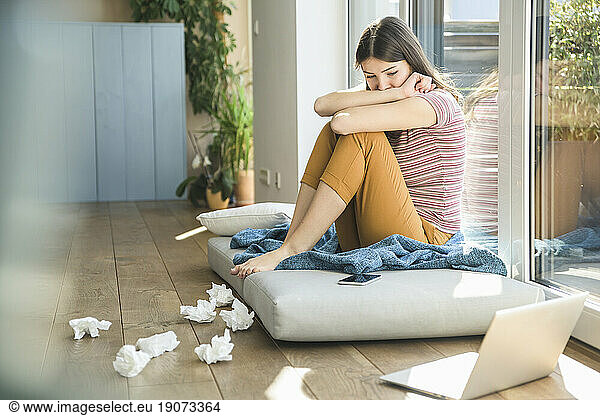 Thoughtful young woman sitting at the window at home with laptop and tissues
