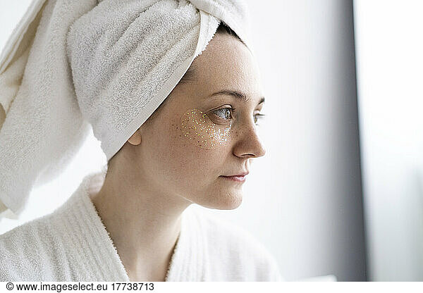 Thoughtful young woman with collagen eye mask wearing towel at home