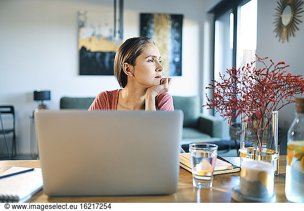 Thoughtful young woman with laptop on desk looking away while sitting at home